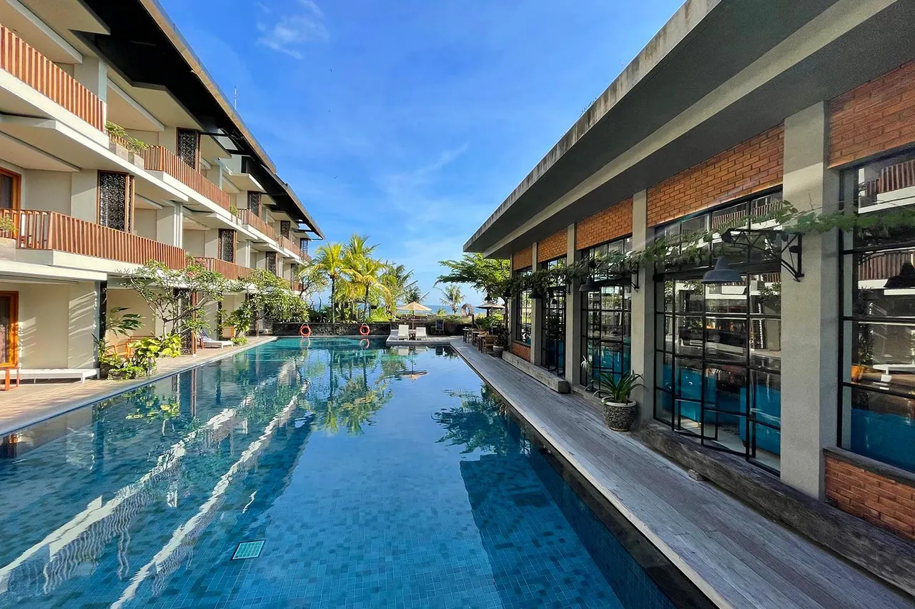 A long lap pool runs between a balconied building on the left and a brick building with tall black-framed windows on the right, with palm trees, sun loungers and an umbrella at the far end under a blue sky.
