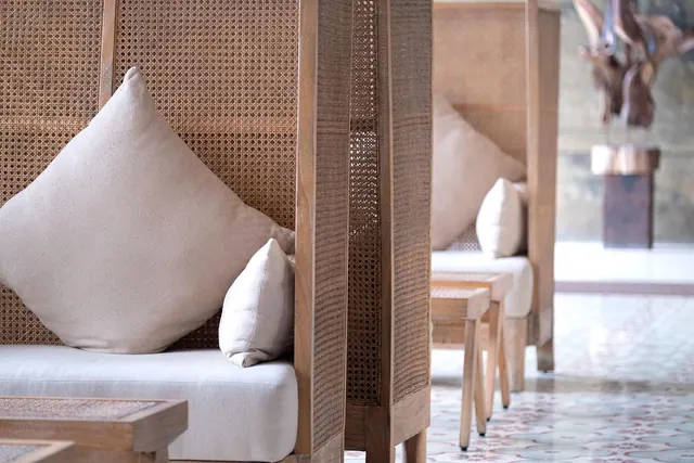 Two high-backed rattan seating booths with cream cushions stand on a patterned tiled floor, with a wooden sculpture in the background.