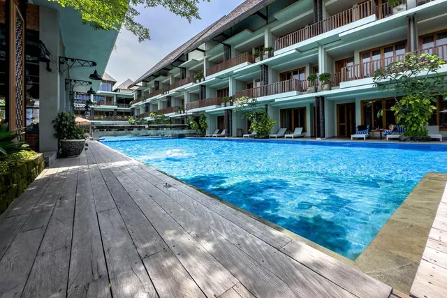 A large blue swimming pool with a timber deck and sun loungers, beside a multi-storey hotel building with balconies.