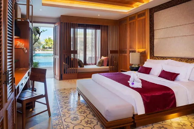 A bedroom with a king bed dressed with a red runner and a towel swan, a wooden desk and chair, a window seat, and an open doorway looking onto a swimming pool and palm trees.