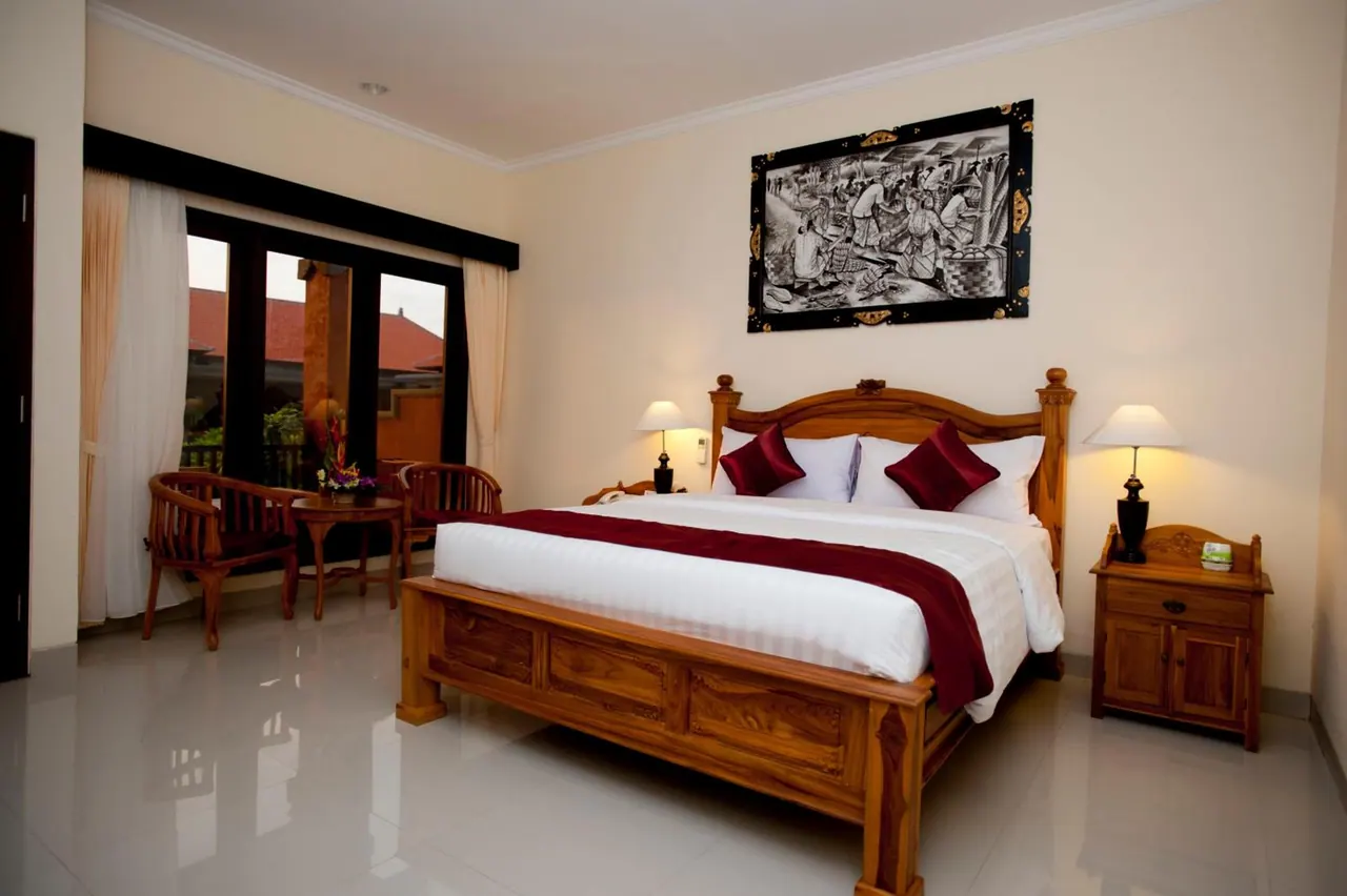 A bedroom with a wooden bed dressed in white and maroon linens, bedside lamps, a framed black-and-white painting, and a window with chairs and a small table beside it.