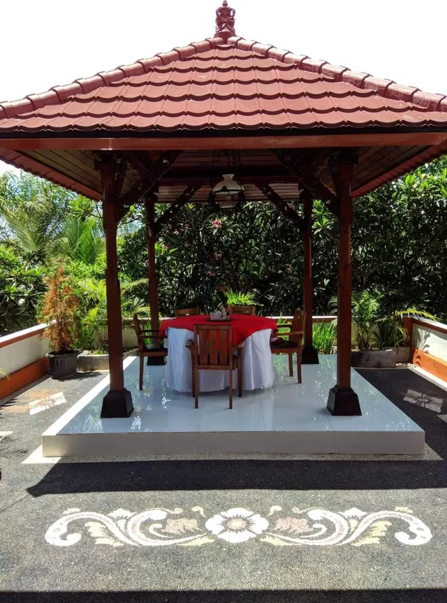 A round dining table with a red and white cloth sits under a red-tiled wooden gazebo surrounded by tropical plants, on a white tiled platform with a patterned paved floor.