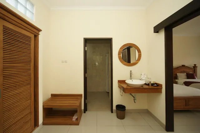 A bathroom with a wall-mounted basin, round mirror, louvred wooden wardrobe and a tiled shower area, with a double bed visible through the doorway on the right.