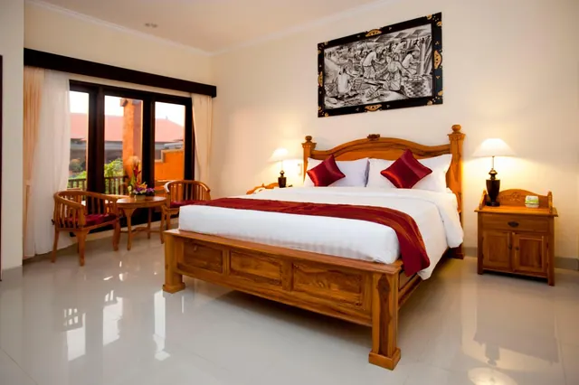 A bedroom with a large wooden bed with red cushions and a red runner, a framed artwork on the wall, bedside lamps, and a small seating area by a window looking out onto roofs and greenery.