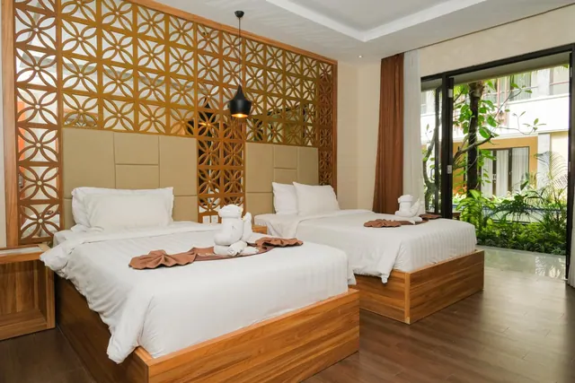 A bedroom with two beds made up with white linen and towel animals, a carved wooden screen behind them, and glass doors opening onto a garden.