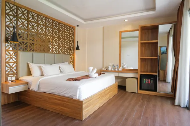 A bedroom with a king bed beneath a carved wooden screen, a mirrored vanity with a kettle, and a wardrobe with a minibar fridge.