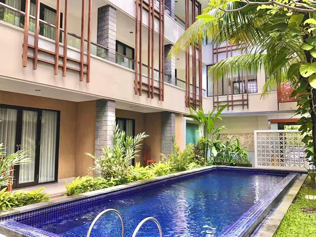 A long blue-tiled swimming pool in a courtyard, lined with tropical plants and backed by a multi-storey building with wooden screens and balconies.