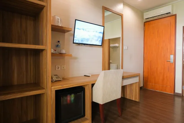 A hotel room corner with a wooden shelf unit, wall-mounted TV, desk with telephone, minibar fridge, chair and an air conditioner above the door.