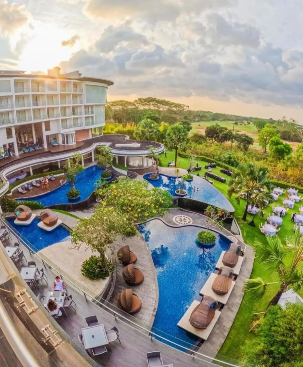 Elevated view of a hotel pool area with curved pools, rattan sun loungers and a lawn with set tables, with trees and a golf course beyond at sunset.