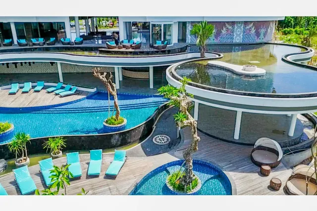 Elevated view of a curved swimming pool, a small round pool with a tree, teal sun loungers on a timber deck, and a lounge area beneath a white building.