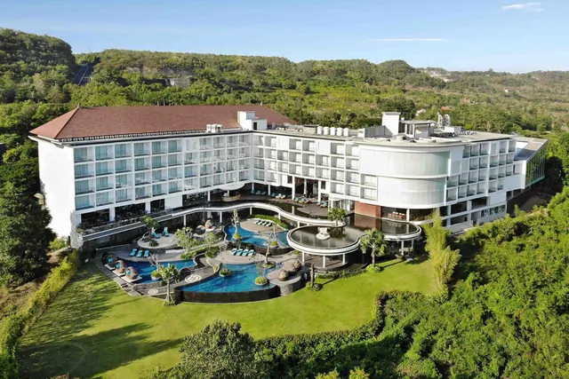 Aerial view of a white multi-storey hotel with curved swimming pools, sun loungers and a lawn, surrounded by forested hills.