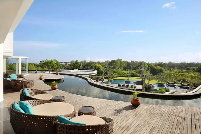 A wooden deck terrace with wicker lounge seats and teal cushions overlooks a winding pool, lawn and tropical trees.