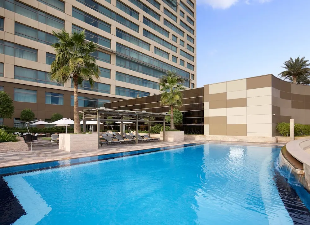A large outdoor pool with a row of sun loungers under a pergola and white umbrellas, beside a multi-storey hotel building.