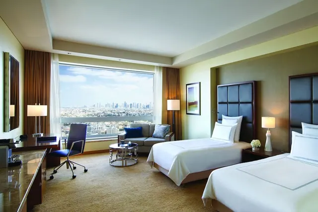 A hotel bedroom with two single beds, a sofa, a desk with a chair, and a large window showing a city skyline.