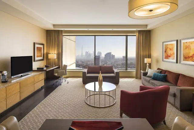 A living room with a sofa, armchairs, coffee table and wall-mounted television, with large windows showing a city skyline including a tall spire.