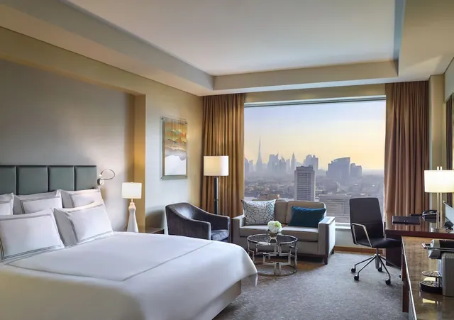 A hotel bedroom with a king bed, a seating area and a desk beside floor-to-ceiling windows, with a city skyline including the Burj Khalifa visible outside.