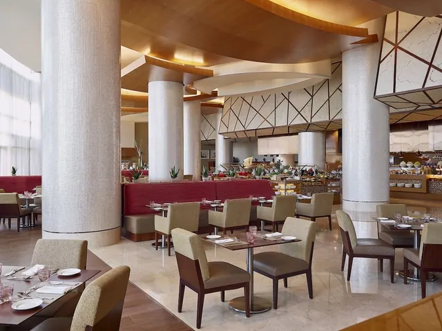 A restaurant dining room with set tables and upholstered chairs, red banquette seating, mosaic-tiled columns, and a buffet station in the background.