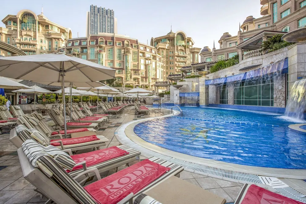 A large curved swimming pool with red-towelled sun loungers and white parasols, surrounded by tall beige buildings.