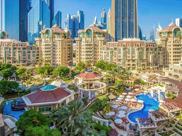 Elevated view of a resort pool surrounded by white umbrellas and sun loungers, with low-rise buildings and a row of tall towers behind.