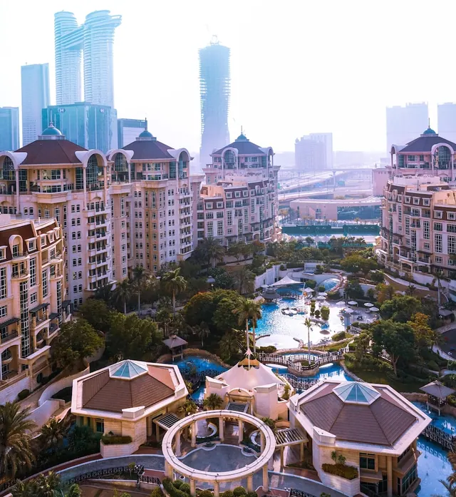 Aerial view of a resort with pools, domed residential-style buildings and a hazy city skyline of towers behind.