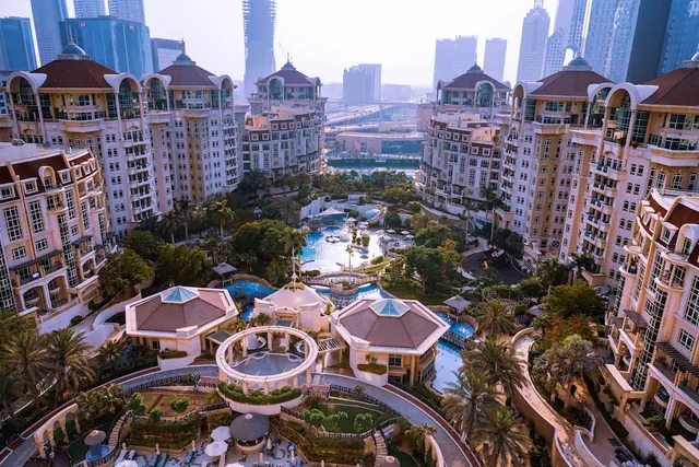 Aerial view of a resort complex with pools, palm trees and low-rise buildings in the foreground, with high-rise towers in the background.