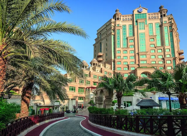 Exterior of a pale pink hotel tower with green-tinted windows, palm trees, and a paved walkway with railings in the foreground under a clear blue sky.