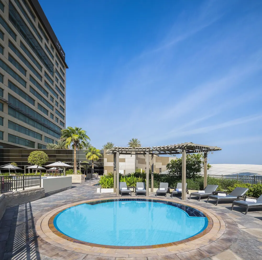 A round outdoor pool with grey sun loungers and a wooden pergola stands beside a tall hotel building under a clear blue sky.