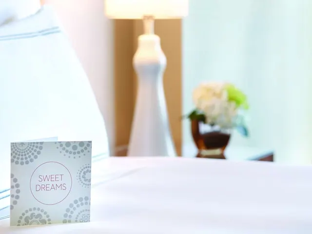 A card reading Sweet Dreams sits on a white bed, with a table lamp and a vase of flowers blurred in the background.