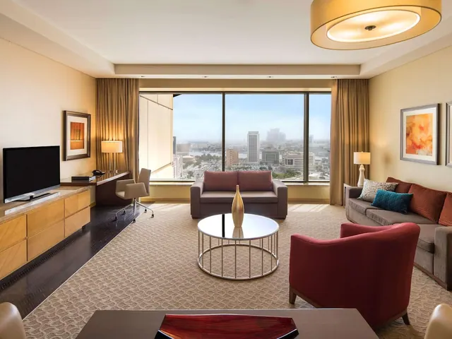 A hotel suite living room with a sofa, armchairs, a round coffee table, a wall-mounted television and a floor-to-ceiling window overlooking a city skyline and water.