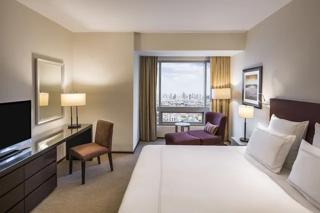 A hotel bedroom with a large bed, a desk with a chair, an armchair by the window, a wall-mounted television, and a view of a city skyline through the window.