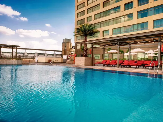 An outdoor swimming pool with red sun loungers and white umbrellas beside a tall hotel building, with a palm tree and a lifebuoy on the deck.