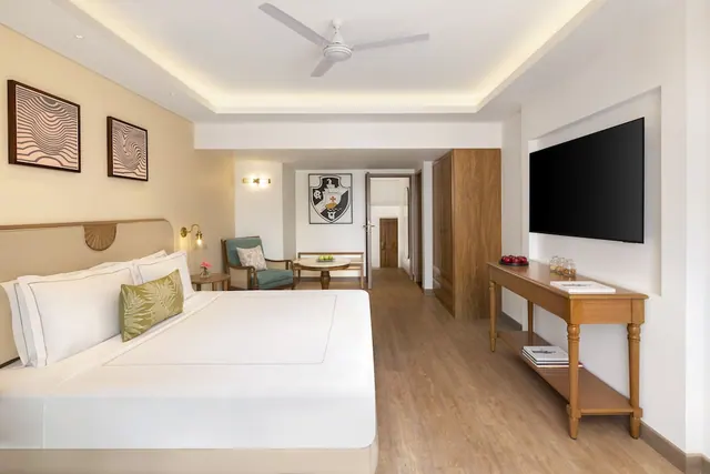 A bright hotel bedroom contains a large bed, seating area, television, wardrobe and ceiling fan.