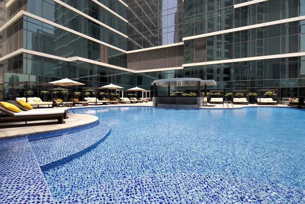 A large mosaic-tiled pool sits in front of a glass-fronted building, with white parasols and cushioned sun loungers along the deck and a covered bar structure in the background.