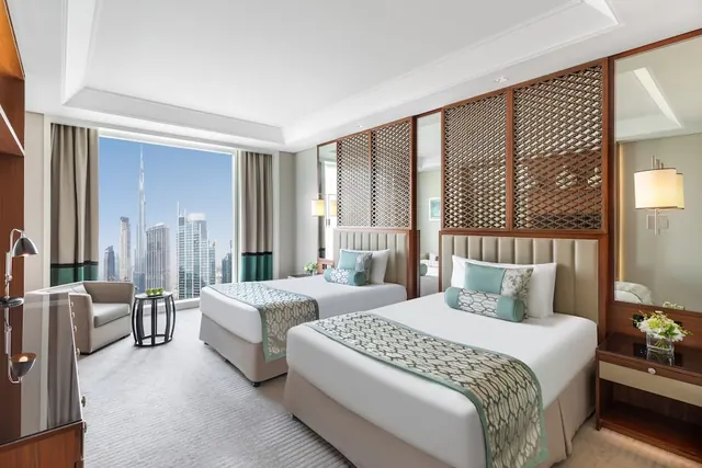 A hotel bedroom with two twin beds and an armchair, with a large window showing the Dubai skyline and the Burj Khalifa.