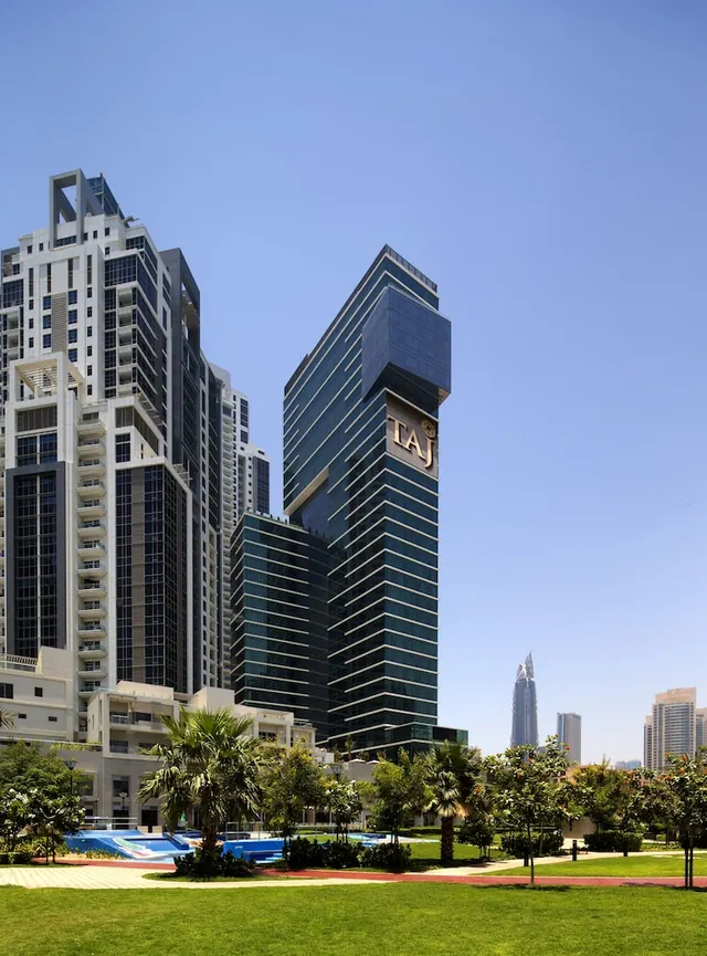 The Taj hotel tower rises above a landscaped lawn, palm trees and a pool under a clear blue sky.