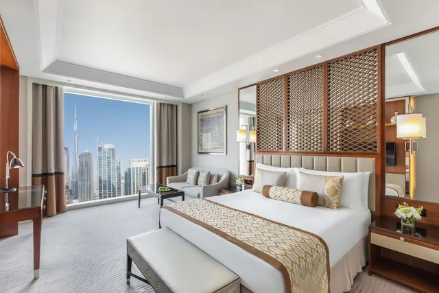A king bed and seating area in a hotel room with floor-to-ceiling windows overlooking a city skyline that includes the Burj Khalifa.