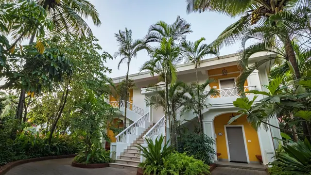 A white-and-yellow hotel building is surrounded by lush tropical plants and palm trees.