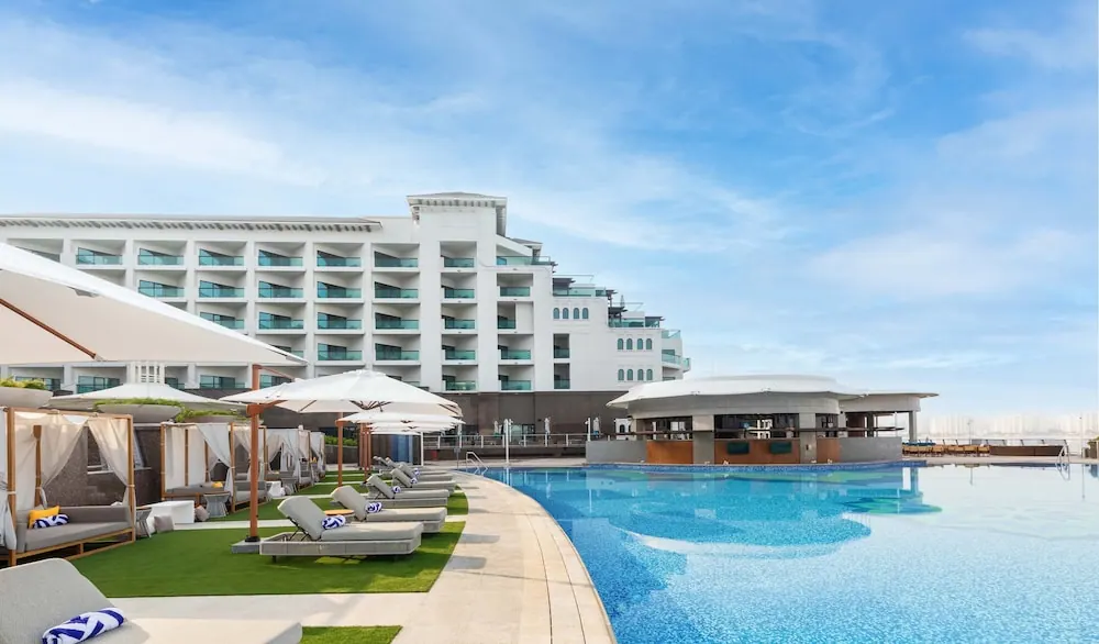 A large pool with sun loungers, cabanas and shade umbrellas stands in front of a white hotel building, with a pool bar to the right.
