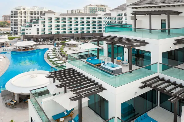 Elevated view from a terrace with a hot tub, blue loungers and a pergola, looking down over a large pool with white umbrellas and sun loungers.
