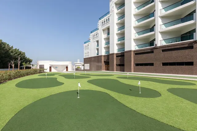 An artificial turf putting green with white flagsticks lies in front of a multi-storey hotel building with glass-balustraded balconies.