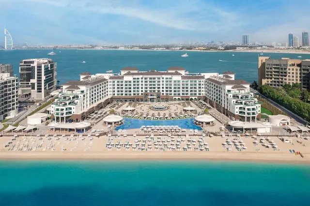 Aerial view of a large beachfront hotel with a curved pool, rows of sun loungers and umbrellas on the sand, and the Burj Al Arab visible across the water.