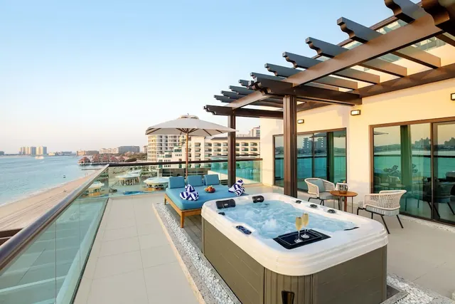 A rooftop terrace with a hot tub, pergola, sun lounger and glass balustrade overlooks the sea and a sandy beach.