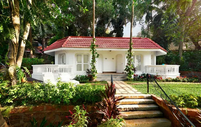 A white heritage-style bungalow with a red tiled roof stands amid tropical gardens.