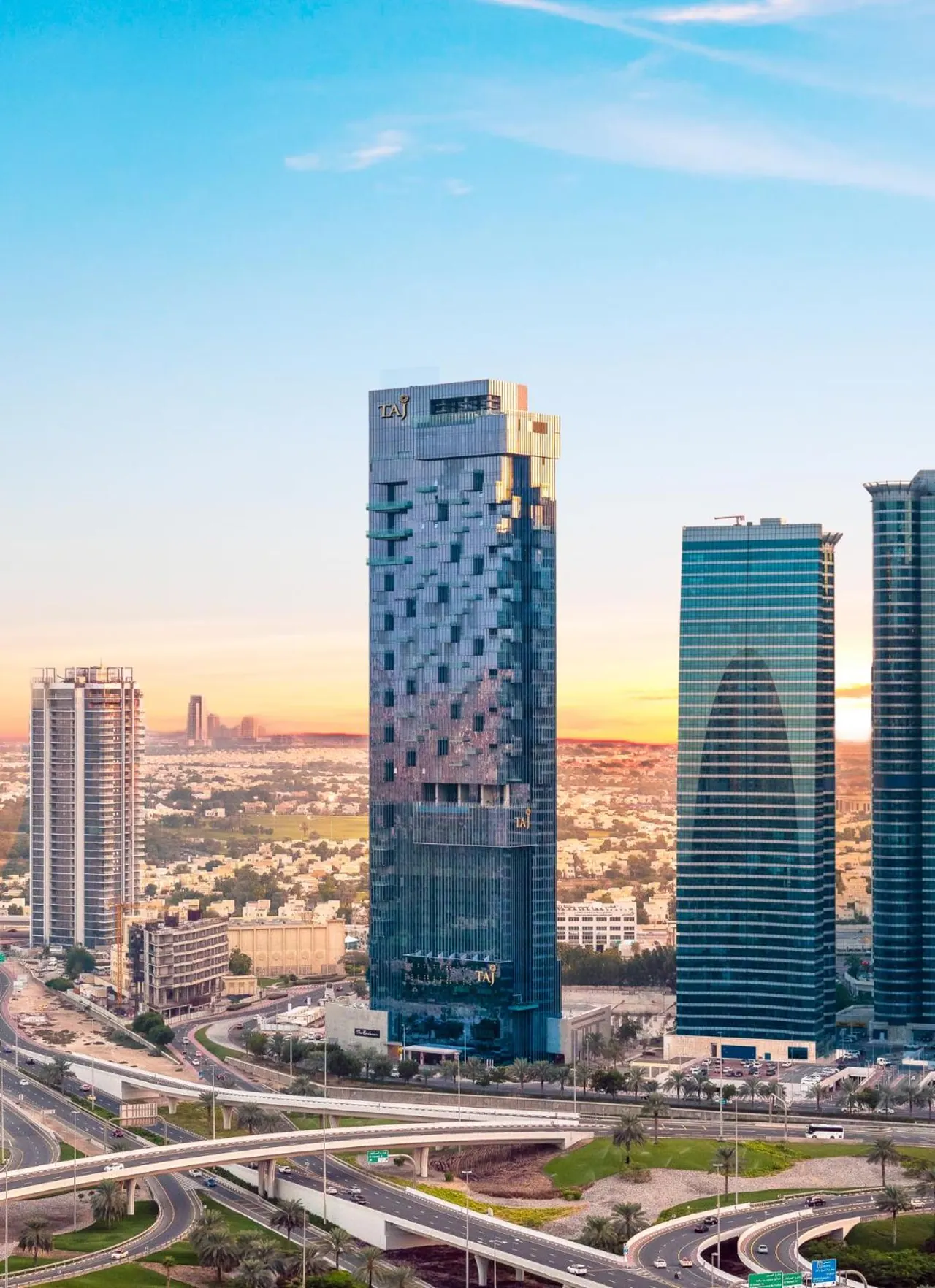 Aerial view of the Taj Dubai tower with surrounding city buildings, highways and palm trees at sunset.
