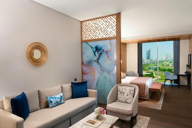 A modern hotel suite living area with a grey sofa, armchair and marble coffee table, with a bed and desk beyond and floor-to-ceiling windows overlooking a city skyline and green landscape.