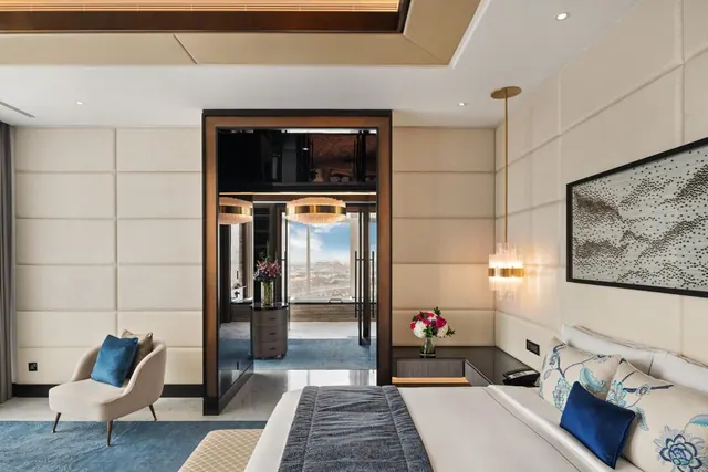 A bedroom with a made king bed, a cream accent armchair with blue cushions, and a doorway through to a room with floor-to-ceiling windows looking out over the city.