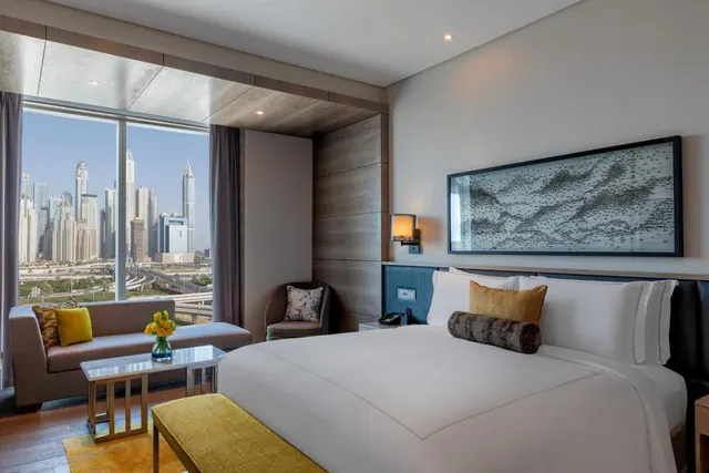 A bedroom with a king bed, a seating area by floor-to-ceiling windows, and a view of high-rise towers outside.