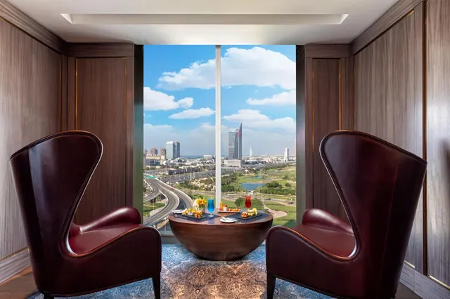 Two leather wingback armchairs and a round wooden table with drinks and snacks face a tall window overlooking a city skyline and green space.