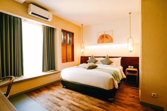 A bedroom with a king bed dressed in white linen and grey cushions, a wall-mounted air conditioner, and a bright window with green curtains.