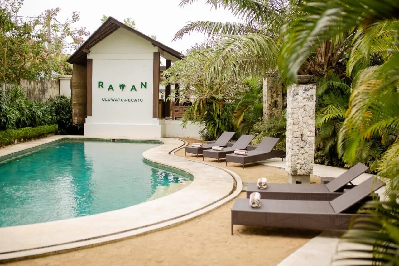 A curved swimming pool with sun loungers and folded towels beside it, with a white building bearing the Ratan Uluwatu, Pecatu sign behind.